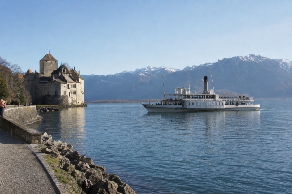 A plain-hulled CGN cruise boat passing Château de Chillon on Lake Geneva near Montreux, mountains behind