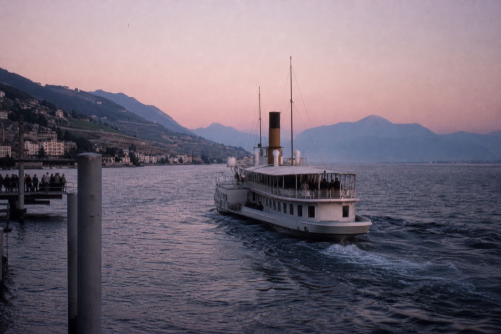 A Belle Époque paddle steamer leaving the Montreux pier on Lake Geneva, the Riviera shoreline and vineyard slopes behind