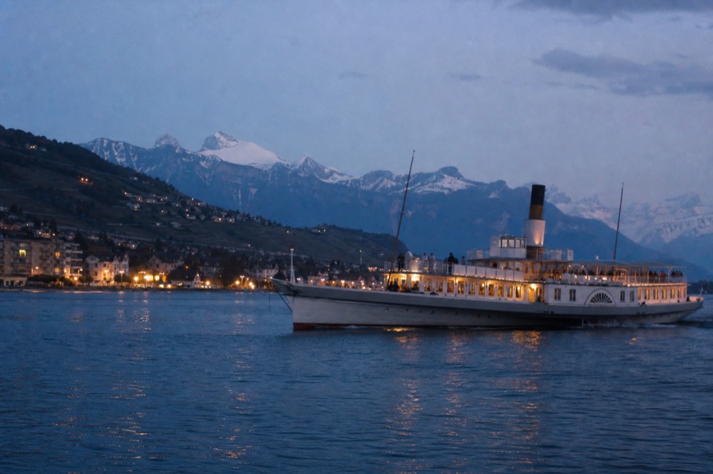 A plain-hull cruise boat on Lac Léman off Lausanne-Ouchy, the Alps across the water and the Lavaux vineyard slopes behind