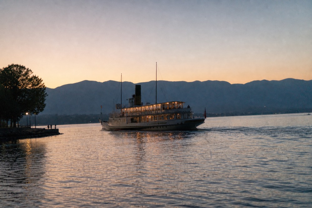 A CGN evening cruise boat on Lake Geneva near Geneva at dusk, warm cabin lights on the water, the Alps a dark line beyond