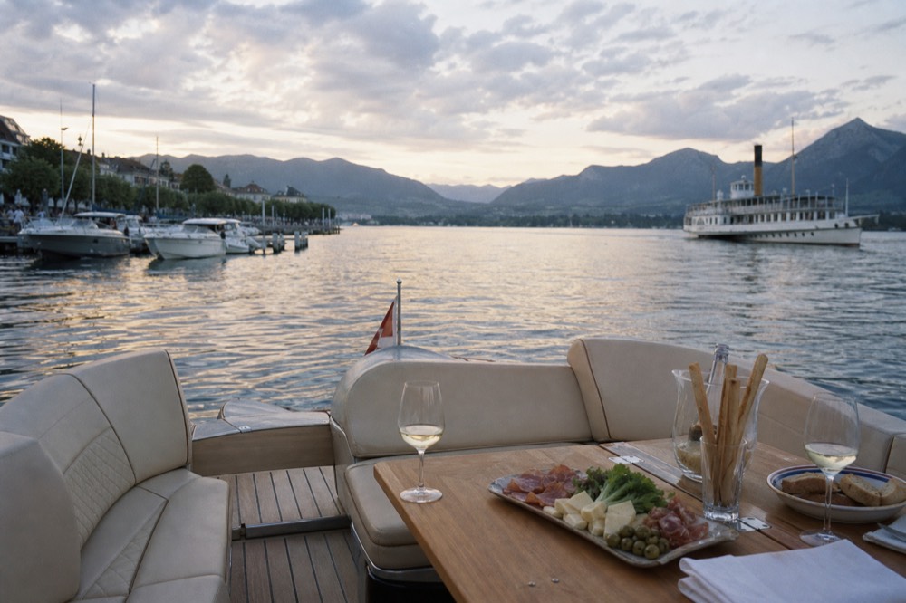 An open private pontoon boat on the Geneva lakefront at golden hour, wine and an aperitif set out on the deck, the harbour behind
