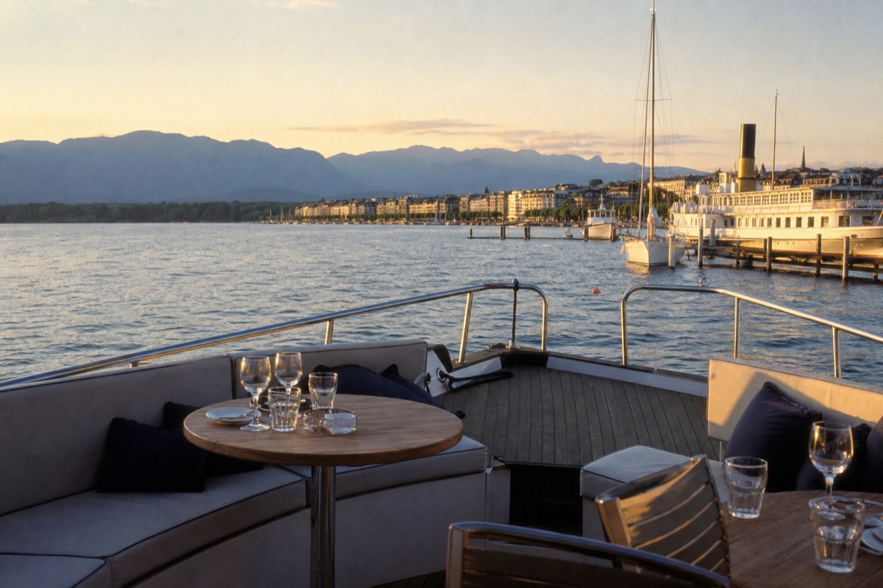 An open pontoon boat on Lake Geneva at golden hour off the Geneva lakefront, glasses and a table set on the deck, the harbour behind