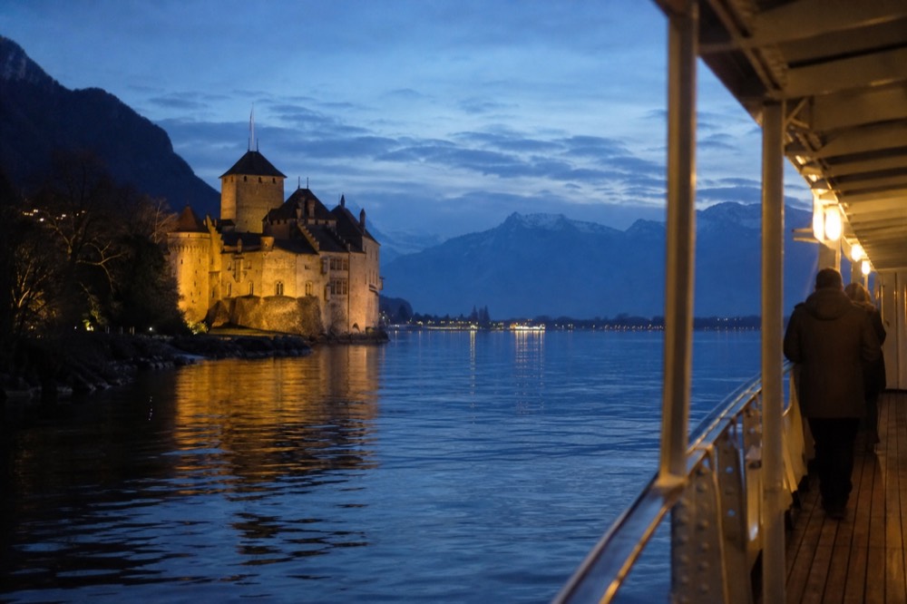 Château de Chillon seen from a CGN boat on Lake Geneva near Montreux, stone towers reflected in calm water with the Alps behind