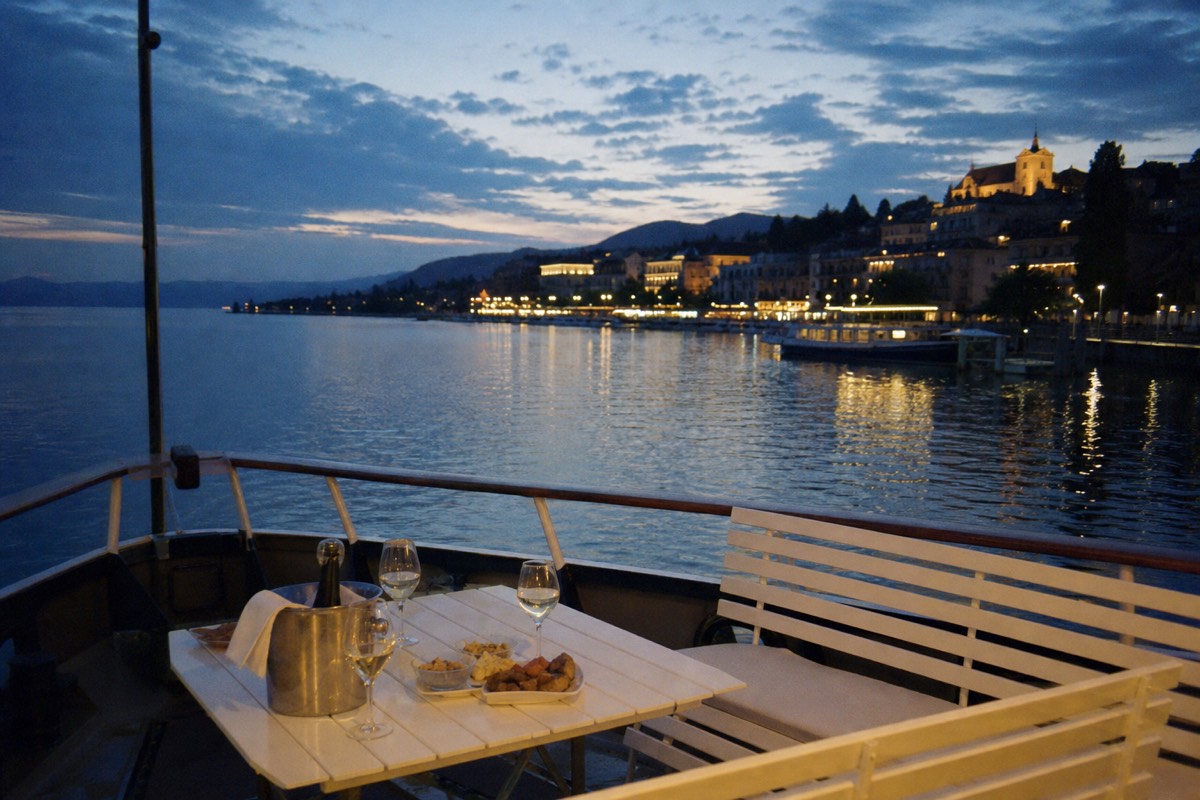 An open pontoon boat on Lake Geneva at dusk, glasses set out on the deck and the shoreline lights coming up behind