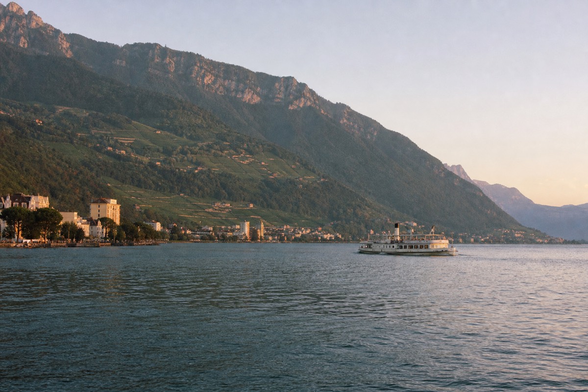 A Riviera cruise boat on Lake Geneva along the Montreux shoreline, vineyard slopes on one side and the Alps across the water
