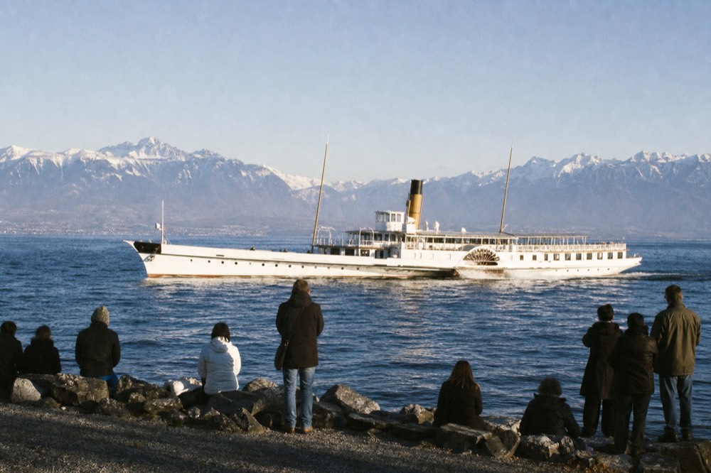 A Belle Époque paddle steamer on Lac Léman off Nyon, its side paddle wheels turning, spectators along the shore