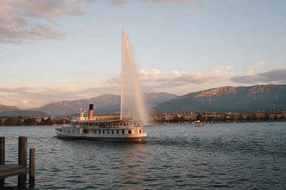 A small cruise boat off the Geneva lakefront on Lake Geneva, the Jet d'Eau throwing water into the air behind it