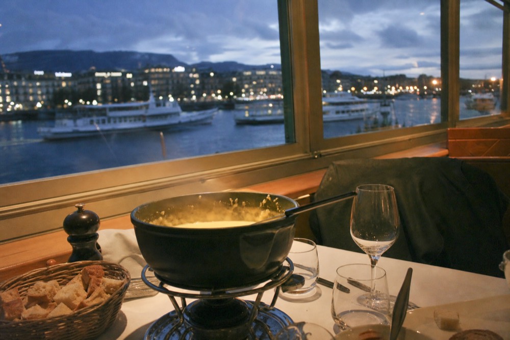 A cheese fondue caquelon on a set table aboard a boat on Lac Léman, Geneva lights through the window at night