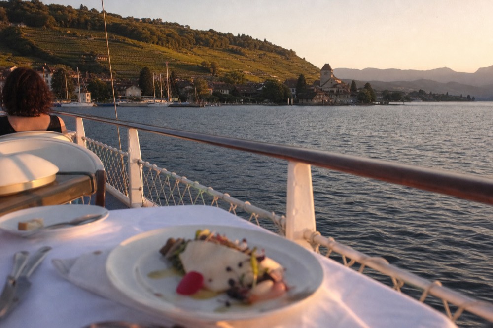 A gourmet plate served on the open deck of a boat on Lac Léman at sunset, the shoreline of Yvoire in the distance
