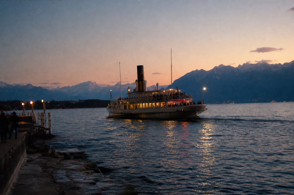 A dining cruise boat on Lac Léman lit at night off the Geneva shore, the Alps a dark line beyond