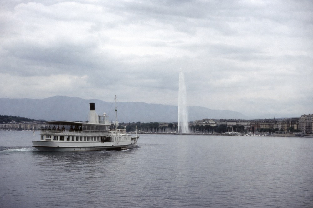 A small cruise boat crossing the Petit Lac off Geneva, the Jet d'Eau and the harbour behind it