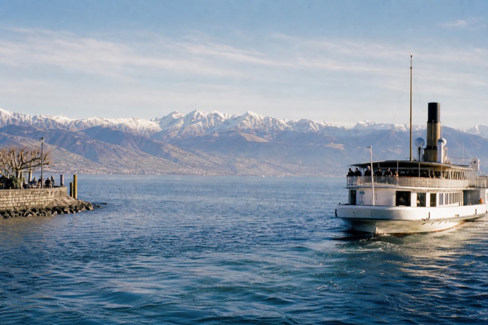 A cruise boat pulling away from the Lausanne-Ouchy waterfront on Lake Geneva with the Alps across the water