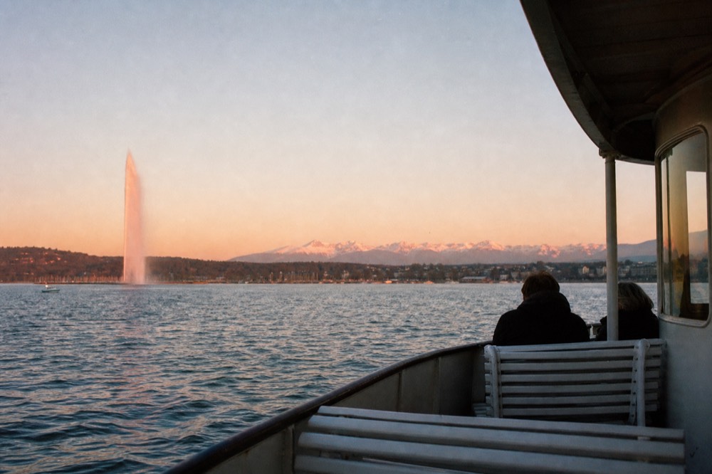 A boat on Lac Léman at sunset off Geneva, the Jet d'Eau catching low golden light and the Alps pink beyond