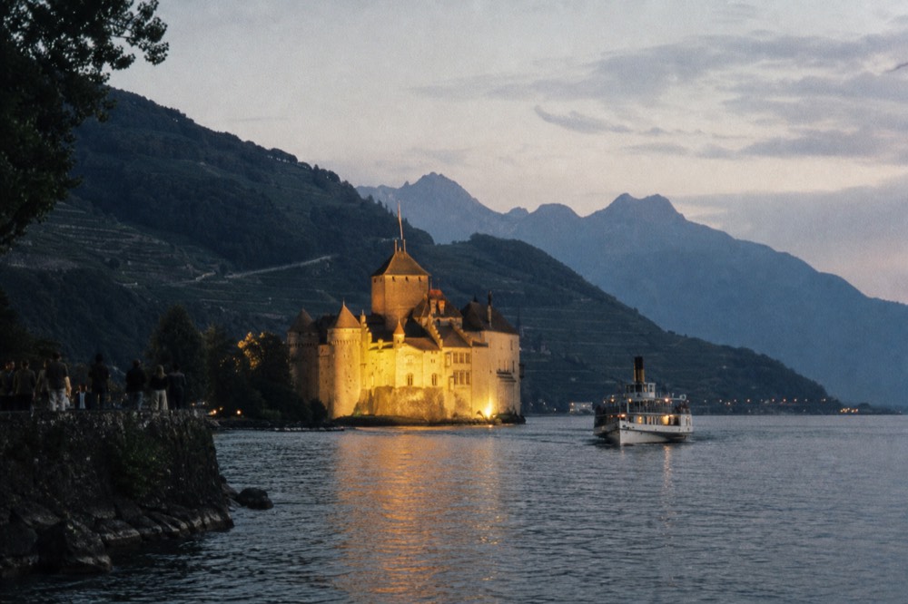 Château de Chillon floodlit at dusk on the shore of Lac Léman, a boat approaching, the Lavaux hills dark behind