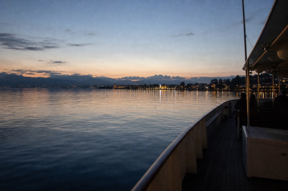 A boat on Lac Léman in the blue hour after sunset off Geneva, shoreline lights coming up along the water
