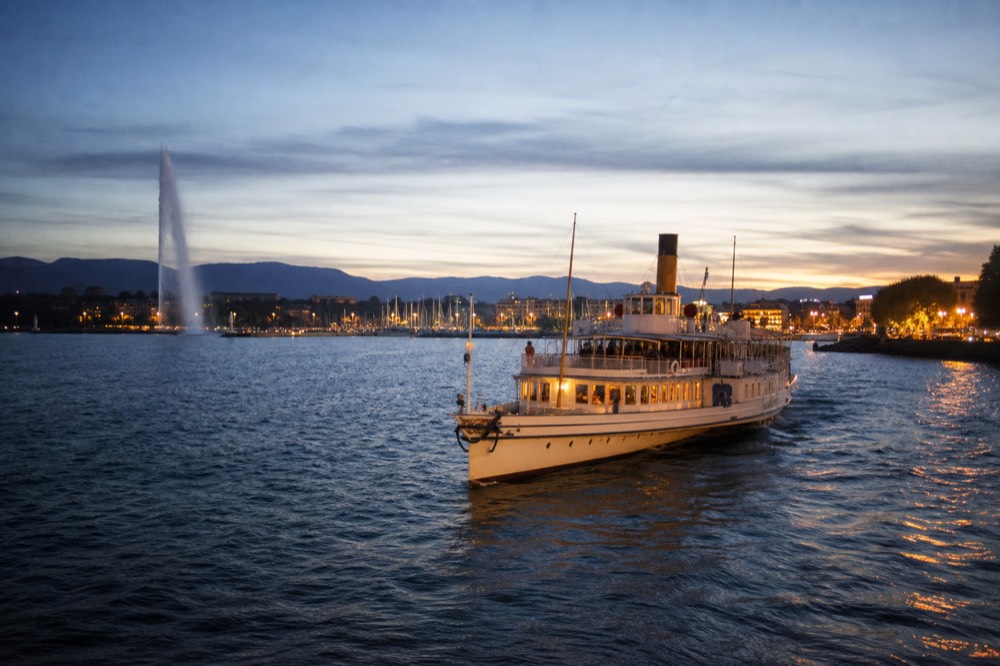 A cruise boat leaving the Geneva lakefront in the evening, the Jet d'Eau and the Rade harbour behind in warm light