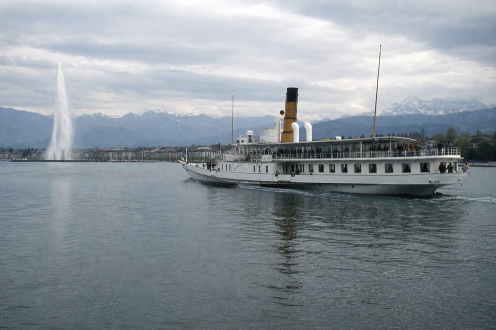 A CGN boat off the Geneva lakefront near the Jet d'Eau, passengers on the open deck and the Alps across Lake Geneva
